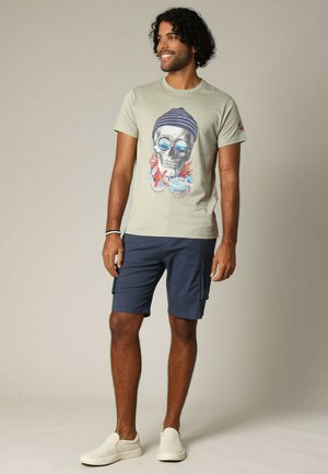 T-shirt vert clair avec un graphique de crâne, associée à un short cargo bleu marine. Le modèle porte des chaussures blanches à enfiler. Tenue décontractée d'été.