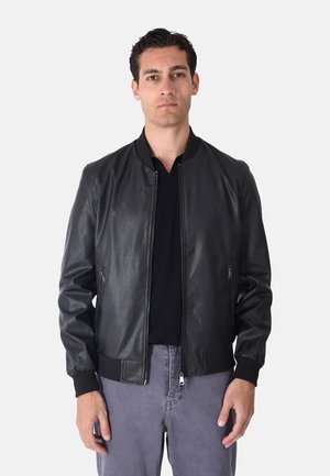 Uomo che indossa una giacca di pelle nera sopra una camicia nera e pantaloni grigi, in piedi davanti a uno sfondo bianco semplice.