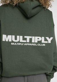 Olivengroene hoodie met een groot wit "MULTIPLY" logo en de tekst "MULTIPLY APPAREL CLUB" op de achterkant; katoenen stof, ontspannen pasvorm.