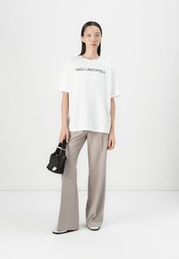 Balta oversize t-shirt su juodu tekstu, smėlio spalvos plačių kojų kelnės ir mažas juodas rankinės. Lygi medžiaga, minimalistinis dizainas.