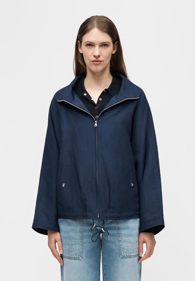 Γυναίκα που φοράει μπλε μπλούζα με φερμουάρ, φαρδιά μανίκια και ρυθμιζόμενο τελείωμα με κορδόνι, συνδυασμένη με ανοιχτόχρωμο τζιν, στέκεται μπροστά σε ένα απλό φόντο.