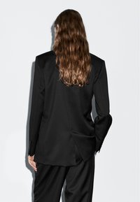 Person mit langen, welligen braunen Haaren steht mit dem Gesicht zur weißen Wand, trägt einen maßgeschneiderten schwarzen Blazer und passende schwarze Hosen.