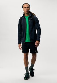 Giacca in pile blu navy, t-shirt verde, pantaloni corti neri con logo, calze grigie e sneakers nere con accenti verdi. Il modello porta uno zaino.