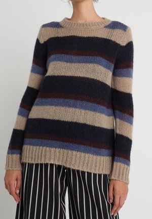 Kobieta ubrana w beżowy sweter w paski w kolorach granatowym, niebieskim i bordowym oraz szerokie spodnie w pionowe, czarno-białe paski na jednolitym tle.