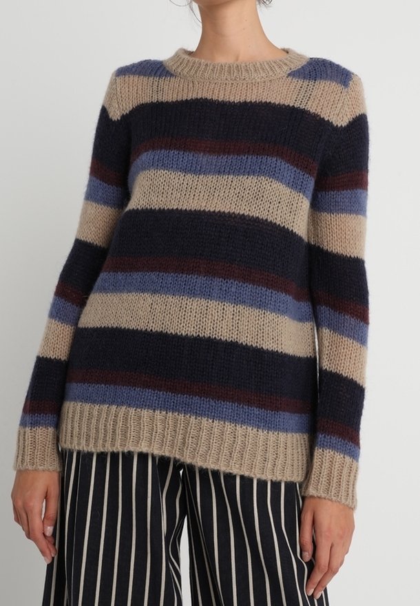 Femme portant un pull en maille rayé beige, marine, bleu et marron, associé à un pantalon large à rayures verticales noires et blanches, sur un fond uni.