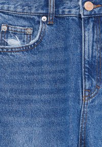 Vaqueros de mezclilla azul claro que presentan un bolsillo desgastado con bordes deshilachados, remaches de cobre y costuras naranjas contrastantes.