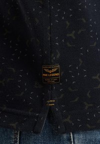 Polo de color marino con tejido texturizado, que presenta un patrón sutil de pequeñas formas en blanco y oliva. Incluye una etiqueta negra con texto dorado.
