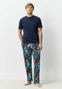 Junger Mann, der barfuß steht, trägt ein marineblaues T-Shirt und bunte Schlafanzughosen mit Blumen- und Elefantenmuster.