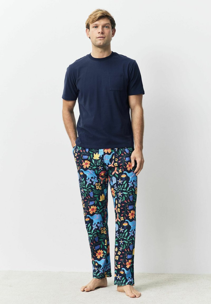 Junger Mann, der barfuß steht, trägt ein marineblaues T-Shirt und bunte Schlafanzughosen mit Blumen- und Elefantenmuster.