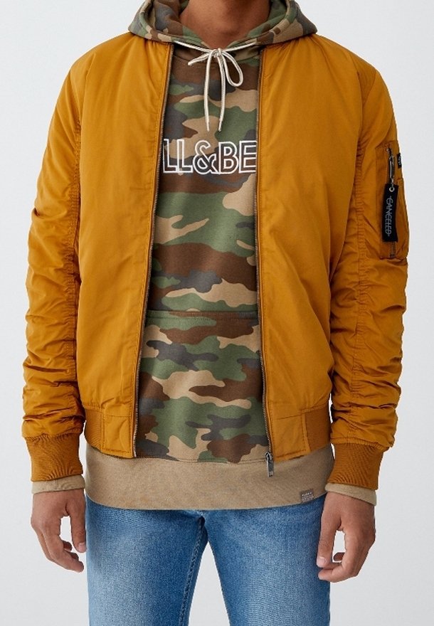 Blouson aviateur en nylon jaune moutarde avec des poignets élastiques, porté sur un sweat à capuche camouflage avec un logo graphique, associé à un jean bleu.