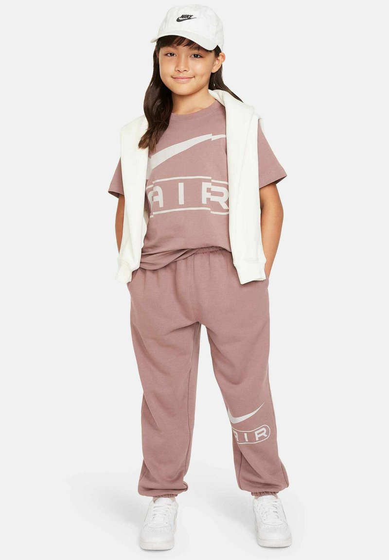 Sudadera rosa claro y pantalones de chándal a juego, hechos de tela suave, con un gran logotipo blanco de Nike en la camiseta, llevados con una gorra blanca.