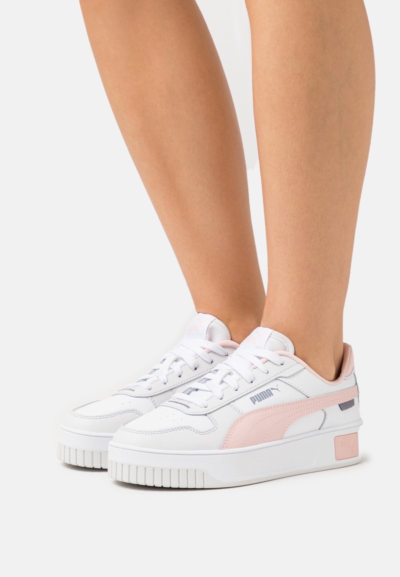 Baskets blanches Puma avec des touches roses portées aux pieds, montrant la vue de côté et une semelle plateforme sur un fond clair uni.
