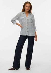 Chemise à boutons avec des rayures verticales bleues et blanches, deux poches poitrine, et les manches retroussées, associée à un jean large foncé et des bottes à talons.