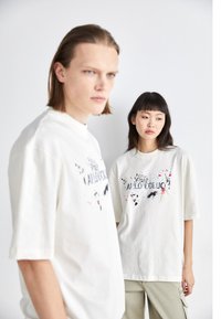 Bomulds oversized t-shirts i off-white med farverigt splattermønster og trykt tekst. Modellen til venstre har korte ærmer; modellen til højre har en afslappet pasform.
