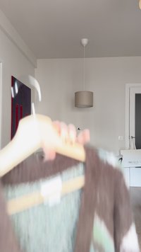 Oseba drži leseni obešalnik z rjavim pletenim puloverjem v minimalistični sobi z belimi stenami, visečo svetilko in vrati v ozadju.