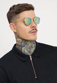 Runda solglasögon med gröna spegelglas, guldmetallram, i kombination med en svart pikétröja med dragkedja och broderad logotyp.