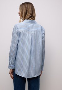 Femme aux cheveux blonds portant une chemise bleu clair à manches longues avec des rayures verticales foncées, vue de dos sur un fond clair uni.