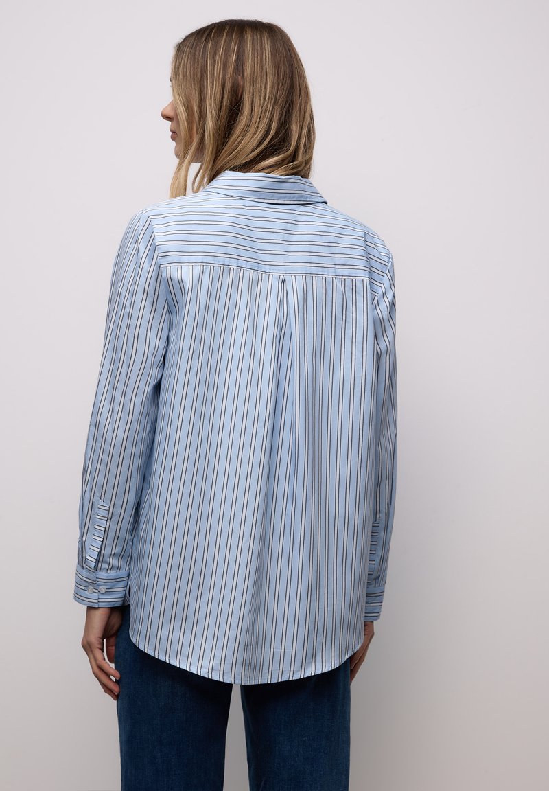 Femme aux cheveux blonds portant une chemise bleu clair à manches longues avec des rayures verticales foncées, vue de dos sur un fond clair uni.