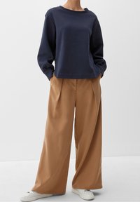 Sweat-shirt bleu marine à col rond et manches longues, associé à un pantalon large couleur beige. Des baskets blanches complètent la tenue.