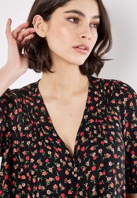 Zwarte bloemenblouse met kleine rode en roze bloemen, een V-hals, geplooid ontwerp en korte mouwen met gatheringdetails.
