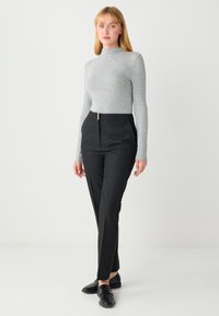 Haut gris à manches longues avec col montant, associé à un pantalon noir taille haute, cintré, avec un pli devant et des détails en métal doré.