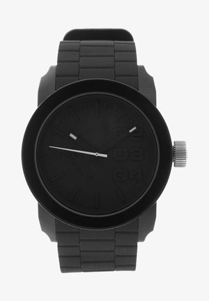 Reloj de pulsera negro con esfera grande redonda, correa negra texturizada, corona plateada a la derecha y marcas sutiles que incluyen los números 2, 3 y 4 en la esfera.
