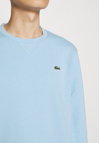 Ljusblå sweatshirt med rundad halsringning, med en liten grön broderad krokodil-logo på vänster bröst. Tillverkad av mjukt tyg.
