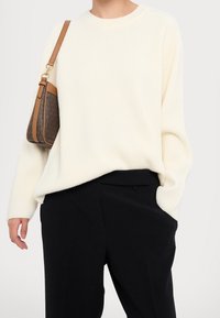 Pull crème côtelé à manches longues, coupe décontractée, associé à un pantalon noir ajusté et un petit sac marron à motif avec une bandoulière beige.