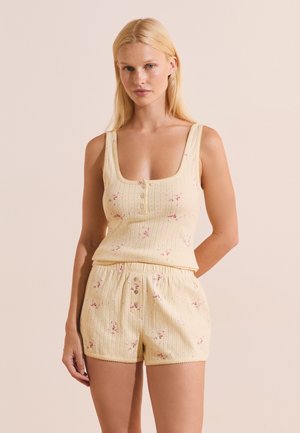 Femme portant un débardeur en maille côtelée jaune pâle et un short assorti avec de petits imprimés floraux roses, debout devant un fond beige.