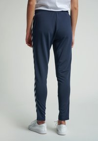 Person som bär marinblå träningsbyxor med vita chevronränder på sidorna och vita sneakers, stående mot en enkel bakgrund.