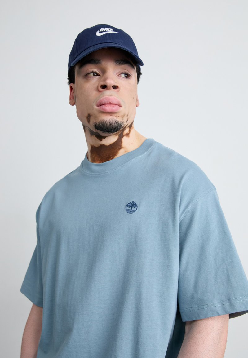 T-shirt en coton bleu à manches courtes et col rond, avec un petit logo d'arbre brodé. Casquette de baseball bleu marine avec le logo Nike blanc.