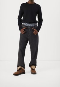 Persona vistiendo una camisa negra de manga larga, jeans oscuros sueltos con la cinturilla de los calzoncillos de cuadros azules visibles, y botas de piel de borrego marrones con la lengüeta afuera.