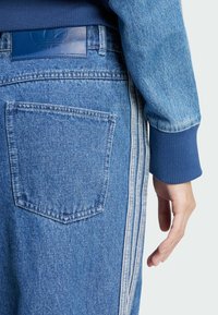 Blue denim pants with a back pocket and a blue label. The side features three vertical white stripes and a ribbed blue cuff at the wrist.