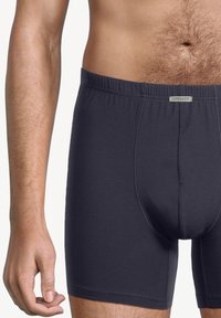 Manlig överkropp i mörkgrå åtsittande kalsonger med en synlig AMMANN-etikett på midjebandet, mot en ljus enfärgad bakgrund.