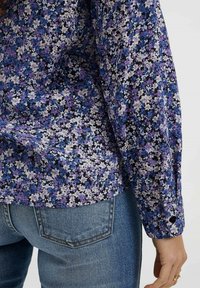 Chemise à imprimé floral avec des fleurs violettes, bleues et blanches sur un fond noir. Dotée d'un poignet avec boutons et d'un tissu doux et léger.