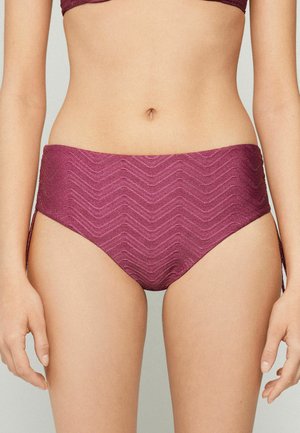 Frau trägt hochgeschnittene, strukturierte lila Bikinihose mit seitlichen Bindebändern, zeigt den Bauchbereich und Hände entspannt an den Seiten.