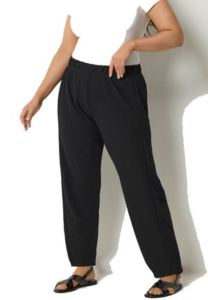 Kobieta nosząca luźne czarne spodnie z elastyczną talią i czarne sandały na paski, pozująca z jedną ręką w kieszeni spodni.