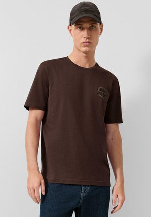 Jonge man draagt een donkerbruine T-shirt met een klein rond logo op de borst en een zwarte pet, staand tegen een effen lichte achtergrond.
