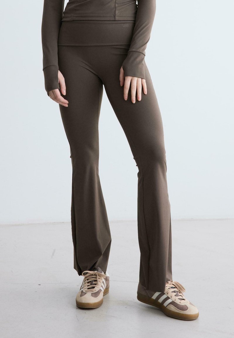 Pantaloni a zampa in colore marrone realizzati in materiale elastico, abbinati a sneaker beige con dettagli marroni. Tessuto liscio, design aderente.