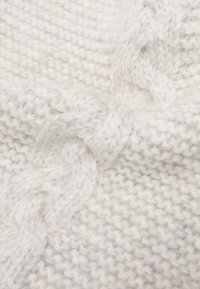 Coperta spessa lavorata a maglia in crema con motivo intrecciato in rilievo e una texture morbida e pelosa. Le cuciture visibili creano un aspetto caldo e accogliente.