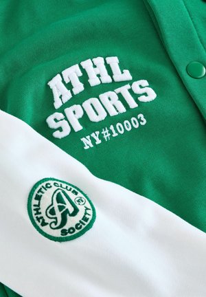 Maillot de sport vert avec des accents blancs. Présente le texte brodé "ATHL SPORTS" et "NY#10003", ainsi qu'un patch logo circulaire sur la manche.