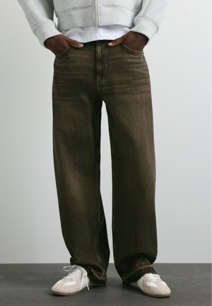 Persona che indossa jeans marrone scuro a gamba larga, felpa con cappuccio grigio chiaro con zip e sneakers bianche, in piedi con le mani nelle tasche.