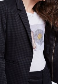 Blazer a scacchi neri con una vestibilità sartoriale, caratterizzato da un unico bottone e rever a punta, indossato su una maglietta bianca con un design grafico.