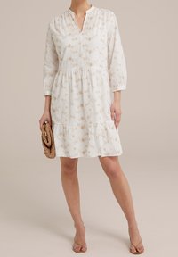 Kvinna klädd i en vit knälång klänning med beige blommönster, hållande en vävd clutch och bärande beige klacksandaler.