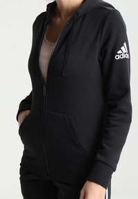 Haut à capuche zippé noir en tissu doux avec une poche avant, des cordons de serrage sur la capuche et un logo Adidas blanc sur la manche gauche.