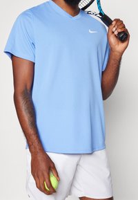 Ljusblå idrottströja med V-ringning, korta ärmar och en liten vit logotyp. Modellen håller en tennisboll och har på sig vita shorts.