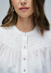 Chemise blanche avec col mandarin, présentant des panneaux en dentelle texturée et des rayures verticales, agrémentée de boutons en forme de perles.