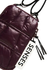 Gepolsterte gesteppte Tasche in tiefburgunderrot, mit Reißverschluss, schwarzem Zubehör und einem weißen Etikett mit dem Wort "SENSES" in fetten Buchstaben.