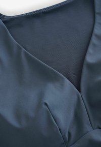 Sötétkék V-nyakú ruha sima anyagból, átlátszó hálós betéttel a nyakvonal alatt, amely textúrakontrasztot mutat.