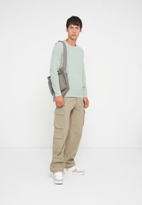 Φωτεινό πράσινο πουκάμισο με μακρύ μανίκι, μπεζ παντελόνι cargo με πλαϊνές τσέπες και λευκά αθλητικά παπούτσια με γκρι λεπτομέρειες. Ανεπίσημος, χαλαρός σχεδιασμός.